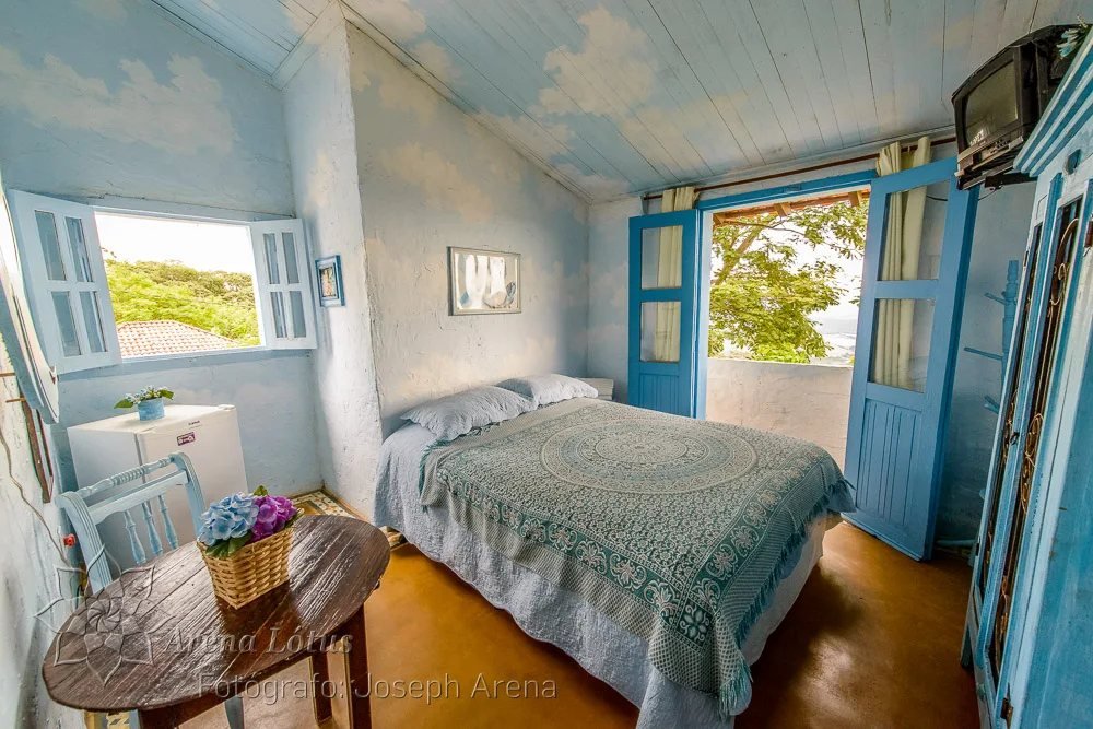 Quarto com teto de nuvens pintadas, colcha estampada e porta-janela azul aberta para a varanda